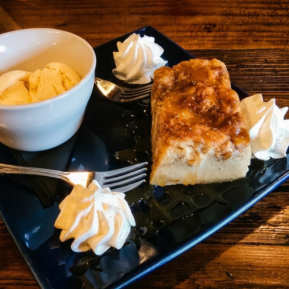Bread pudding with caramel sauce, whipped cream, and a side of vanilla ice cream on a wooden table.